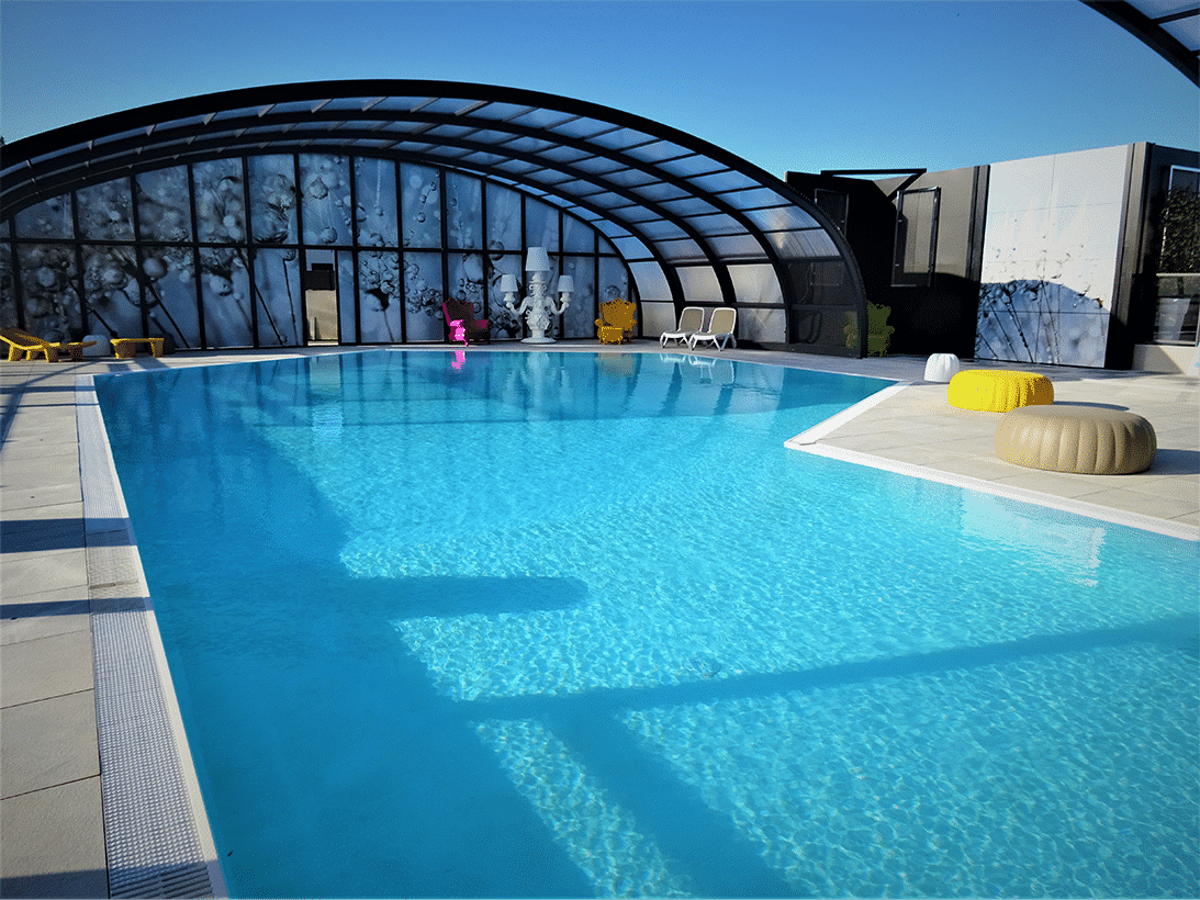 Bassin de piscine turquoise sous verrière arrondie, transats et poufs jaunes au bord